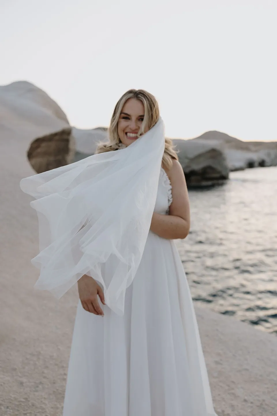 Georgina smiling beside an inlet with her veil held over her shoulder, at Sarakiniko on Milos in the Cyclades, Greece, during an Elopement Collective elopement. Photograph by George Bowden.