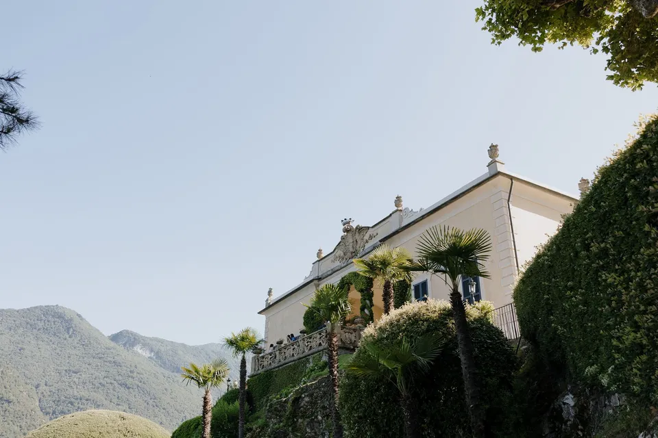 Villa del Balbianello’s pale facade, stone parapet and garden hedges seen from below against a blue sky, at Lake Como in Lombardy, Italy, for an Elopement Collective elopement. Photograph by George Bowden.