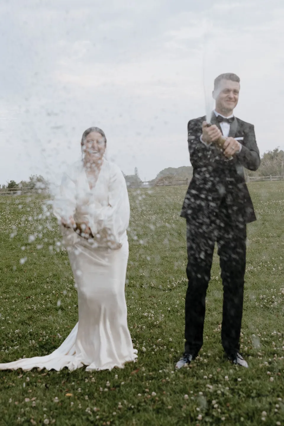 Laura and Matthew laughing as sparkling wine sprays through the air. At The Farm and Three Blue Ducks in Ewingsdale, Byron Bay, for an Elopement Collective elopement. Photograph by George Bowden.