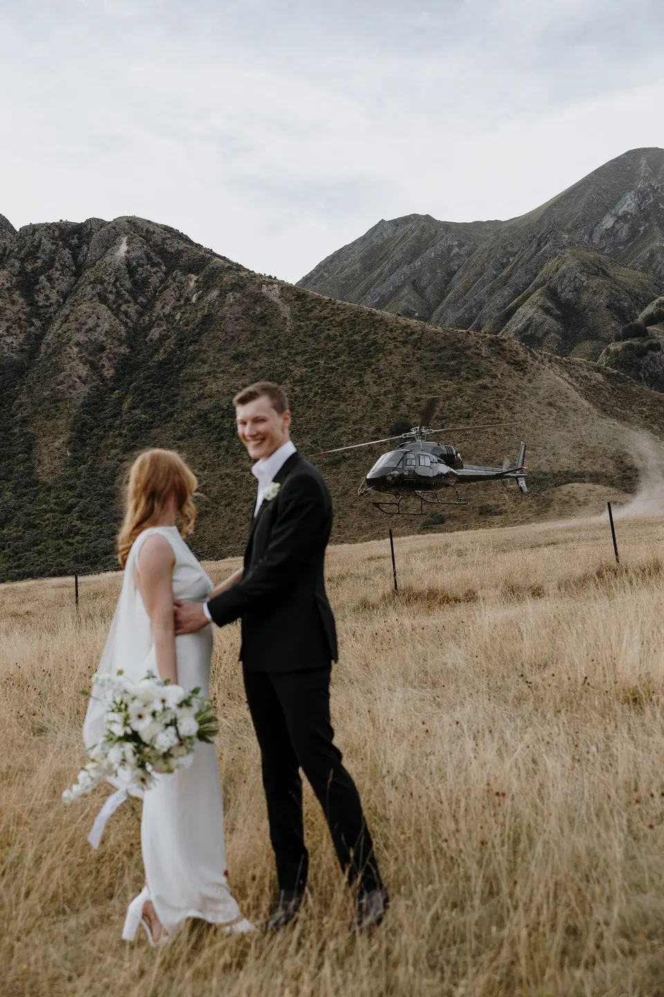 Neve holding her white Orange Blossom bouquet as she stands with Hamish in the grass, with their helicopter behind them, at Moke Lake in Queenstown, New Zealand, for an Elopement Collective Queenstown elopement. Photo by George Bowden.