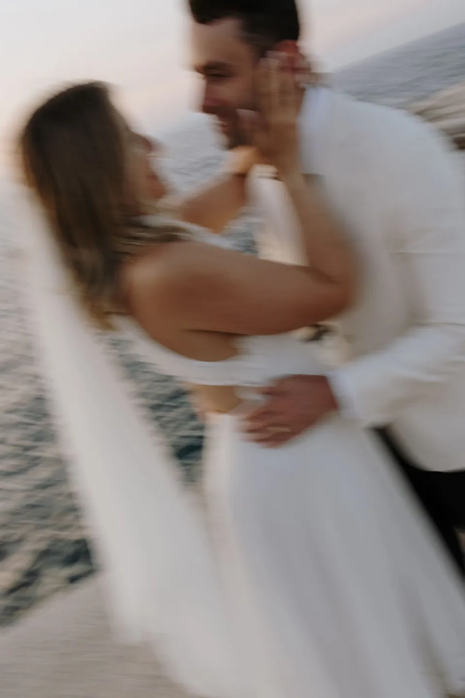 A portrait with intentional motion blur of Georgina and Brad smiling and embracing, with her hand against his face, at Sarakiniko on Milos in the Cyclades, Greece, during an Elopement Collective elopement. Photograph by George Bowden.