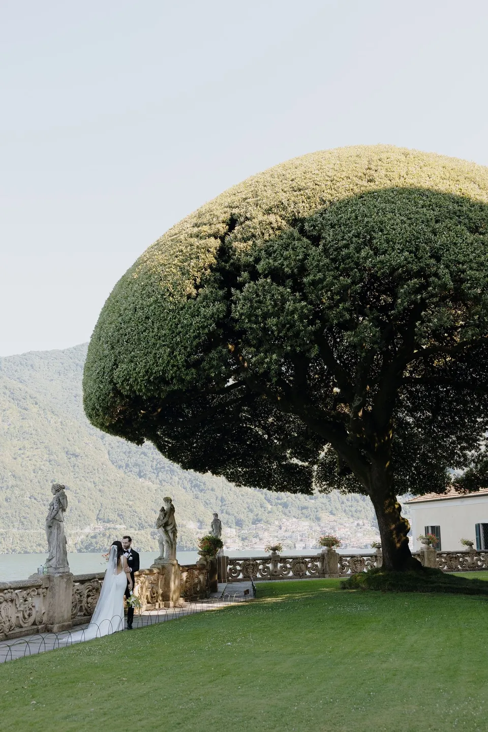 Lyndal and Lachlan standing beside a sculpted tree and garden balustrade at Villa del Balbianello above the lake, at Lake Como in Lombardy, Italy, for an Elopement Collective elopement. Photograph by George Bowden.