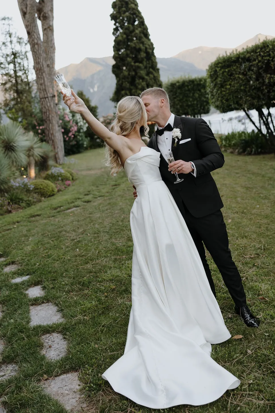 Zoe raising a champagne flute as Charlton stands beside her on the lakeside lawn, at Passalacqua in Moltrasio, Lake Como, Italy, for an Elopement Collective elopement. Photo by George Bowden.