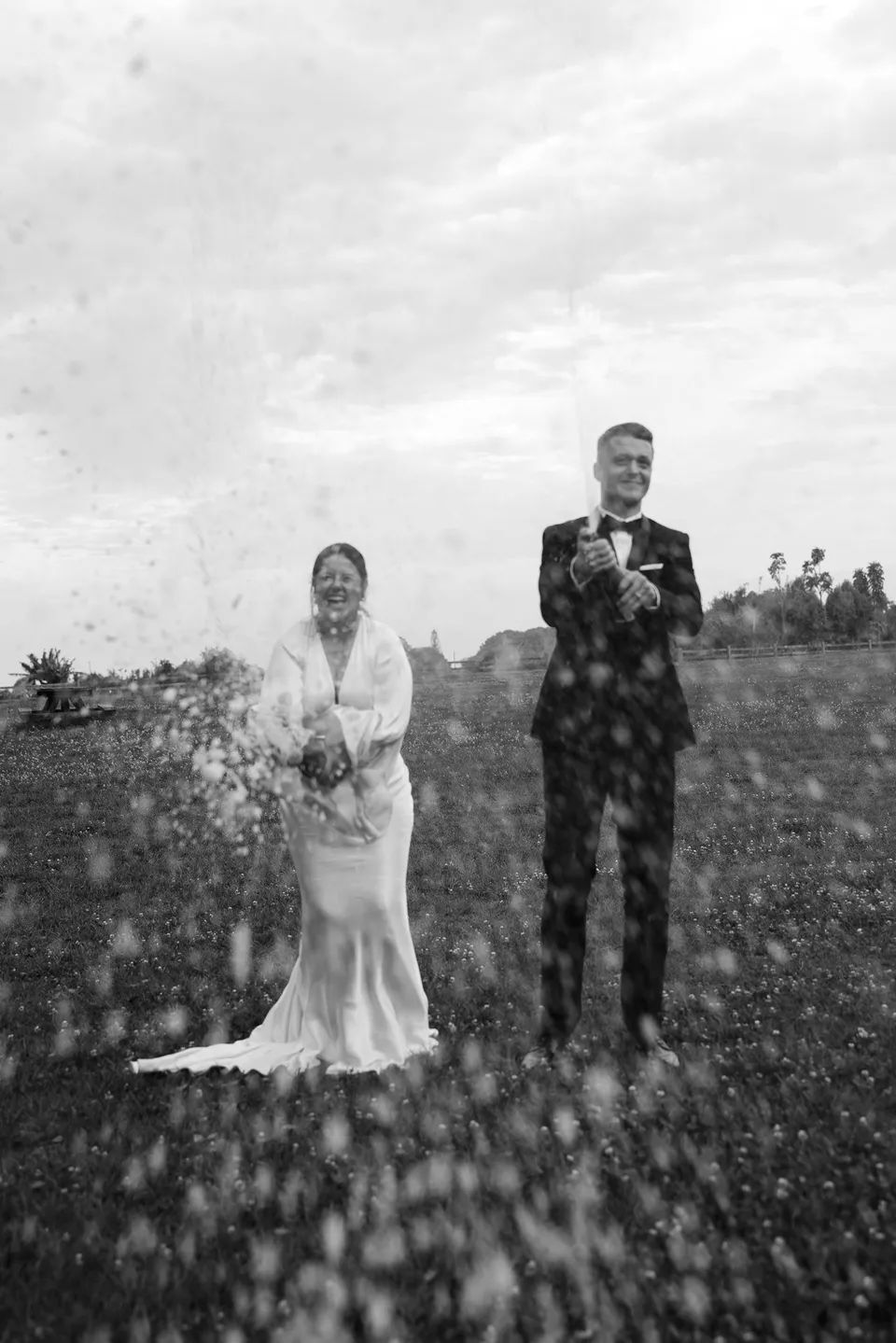 Black-and-white photograph of Laura and Matthew celebrating with a spray of sparkling wine. At The Farm and Three Blue Ducks in Ewingsdale, Byron Bay, for an Elopement Collective elopement. Photograph by George Bowden.