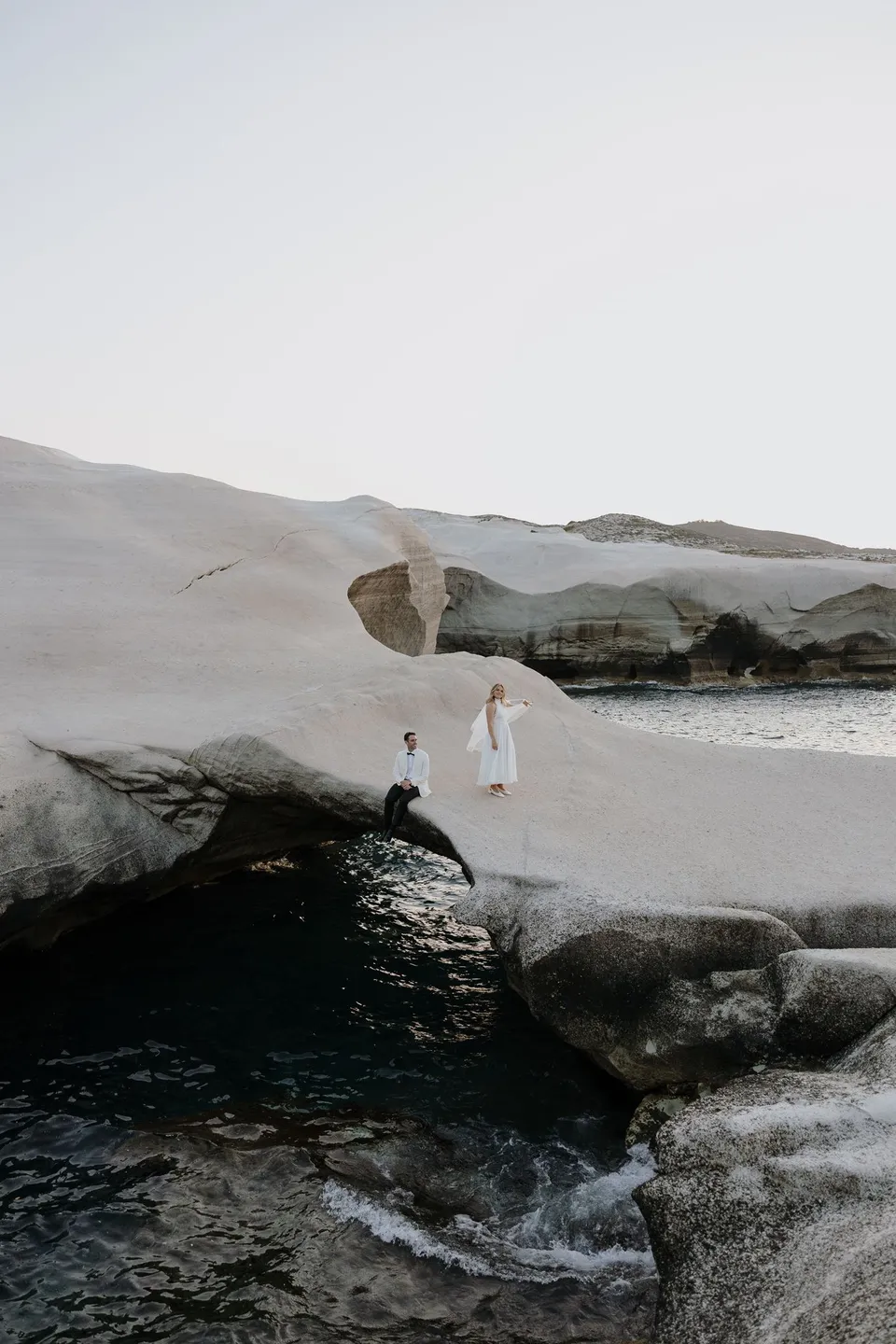 Georgina and Brad holding hands as they step across low rocks beside the water, with the white coastline behind them, at Sarakiniko on Milos in the Cyclades, Greece, during an Elopement Collective elopement. Photograph by George Bowden.