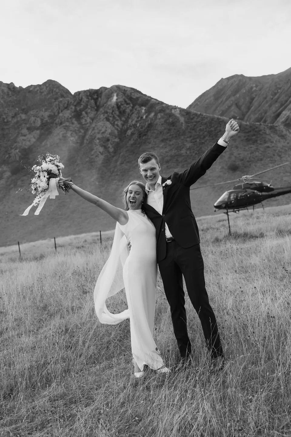 A black and white portrait of Neve and Hamish raising their arms in celebration, with Neve holding her Orange Blossom bouquet and a helicopter behind them, at Moke Lake in Queenstown, New Zealand, for an Elopement Collective Queenstown elopement. Photo by George Bowden.