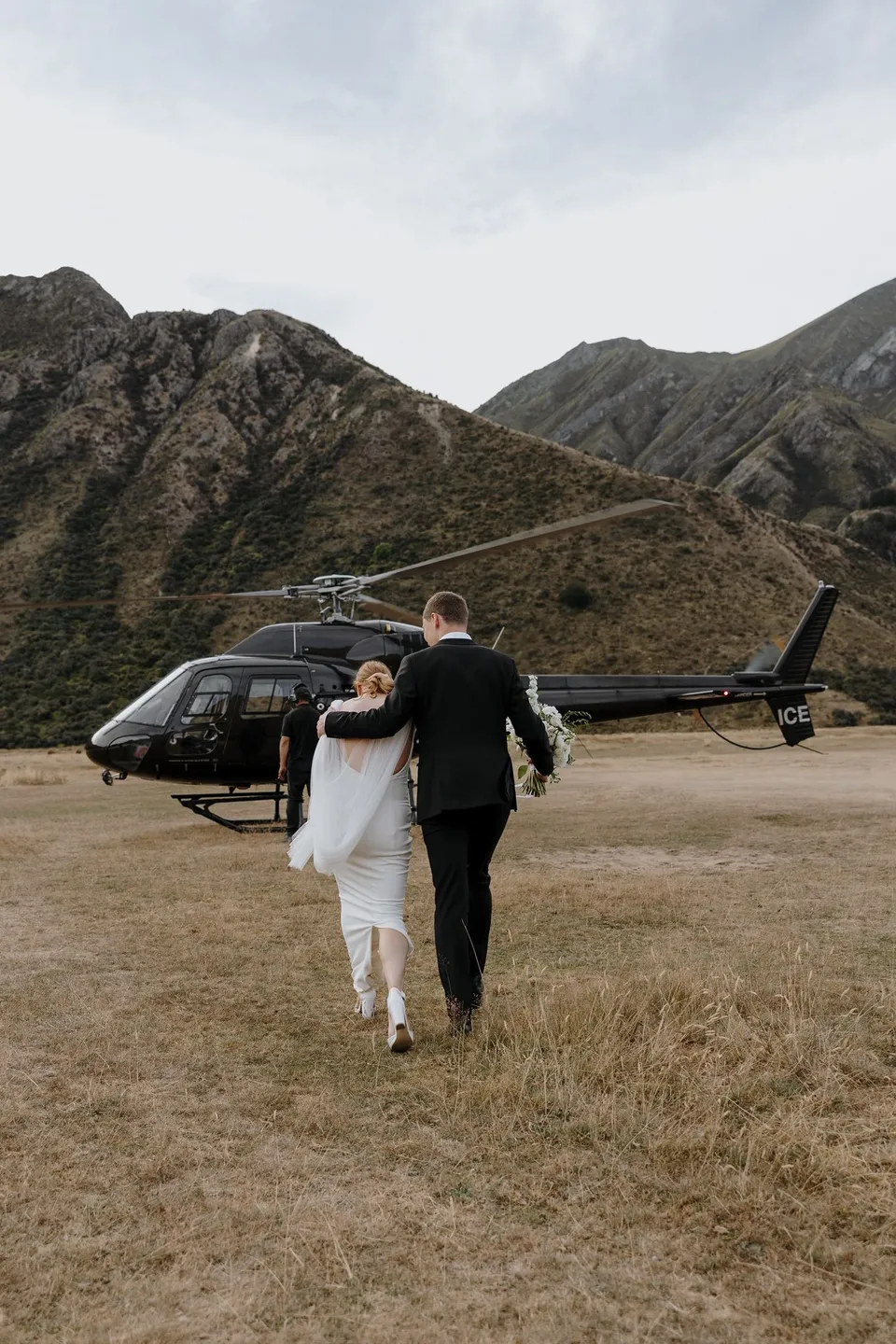 Neve and Hamish walking arm in arm across the grass towards their helicopter for mountain portraits, at Moke Lake in Queenstown, New Zealand, for an Elopement Collective Queenstown elopement. Photo by George Bowden.