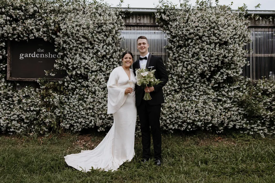 Laura and Matthew standing together beside a flowering wall at The Farm. At The Farm and Three Blue Ducks in Ewingsdale, Byron Bay, for an Elopement Collective elopement. Photograph by George Bowden.