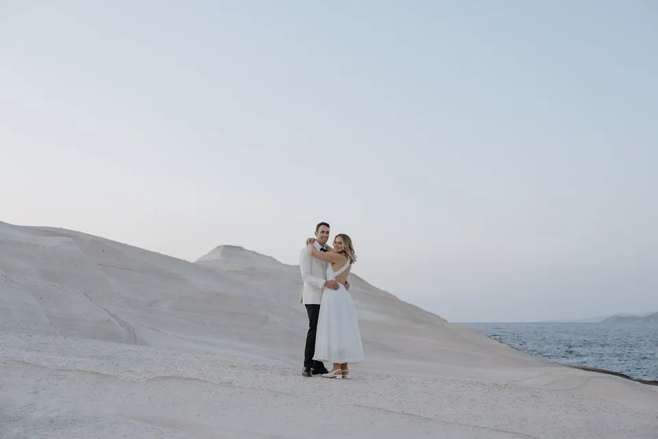 Georgina and Brad embracing among pale coastal rocks beneath a soft evening sky, at Sarakiniko on Milos in the Cyclades, Greece, during an Elopement Collective elopement. Photograph by George Bowden.
