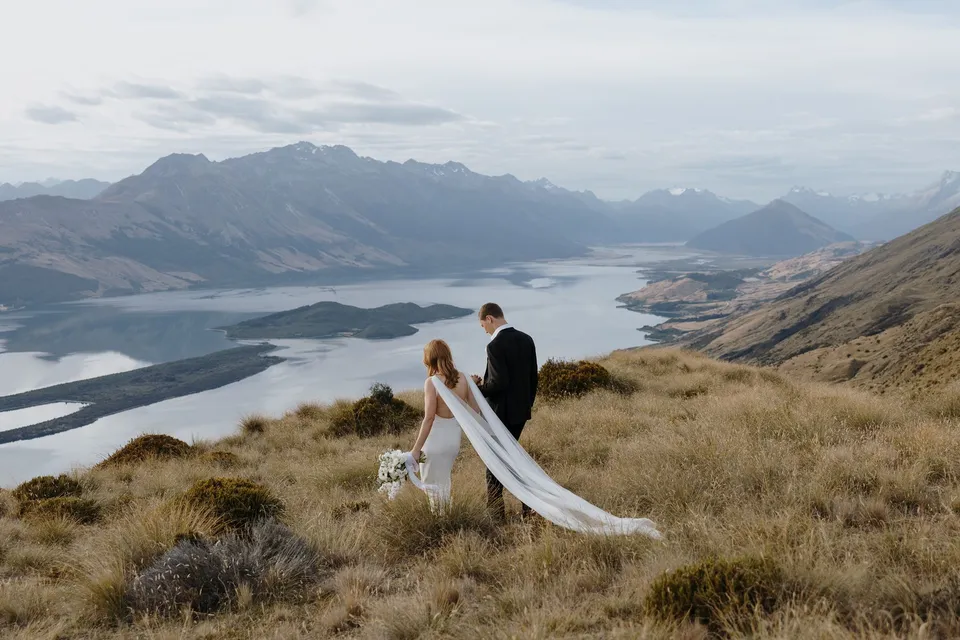 Neve and Hamish with a white Orange Blossom bouquet and flowing cape above Lake Wakatipu, Pig Island and Pigeon Island, with views towards Glenorchy, on Big Geordie above Queenstown, New Zealand, for an Elopement Collective Queenstown elopement. Photo by George Bowden.