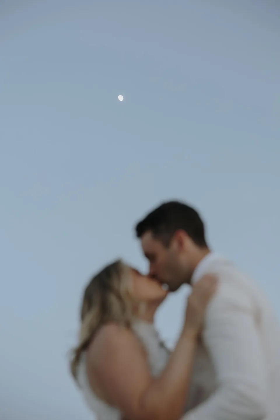 A soft-focus portrait of Georgina and Brad kissing beneath a small moon in the blue evening sky, at Sarakiniko on Milos in the Cyclades, Greece, during an Elopement Collective elopement. Photograph by George Bowden.
