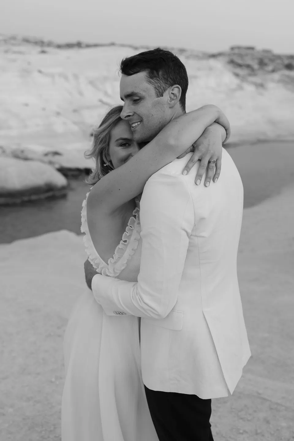 Black and white photograph of Georgina hugging Brad around his shoulders as he smiles, at Sarakiniko on Milos in the Cyclades, Greece, during an Elopement Collective elopement. Photograph by George Bowden.