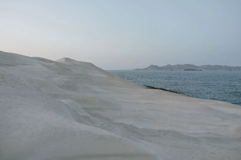 Pale coastal rock slopes beside the sea, with distant island hills beneath an evening sky, at Sarakiniko on Milos in the Cyclades, Greece, during an Elopement Collective elopement. Photograph by George Bowden.