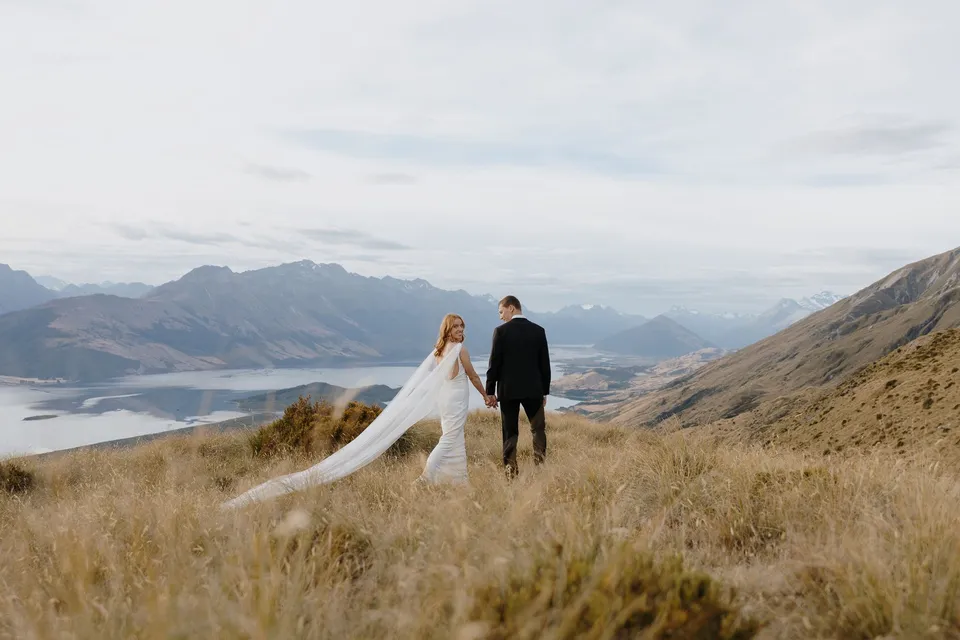 Neve and Hamish walking together through golden tussock high above Lake Wakatipu, with mountains stretching into the distance, on Big Geordie above Queenstown, New Zealand, for an Elopement Collective Queenstown elopement. Photo by George Bowden.