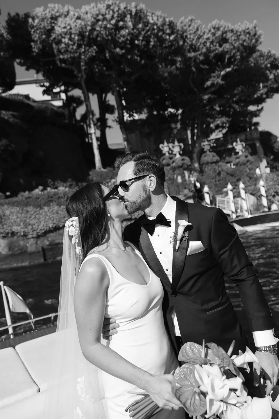 Black and white photograph of Lyndal and Lachlan kissing aboard a boat, wearing sunglasses with her white bouquet by Garden of Love in the foreground, at Lake Como in Lombardy, Italy, for an Elopement Collective elopement. Photograph by George Bowden.
