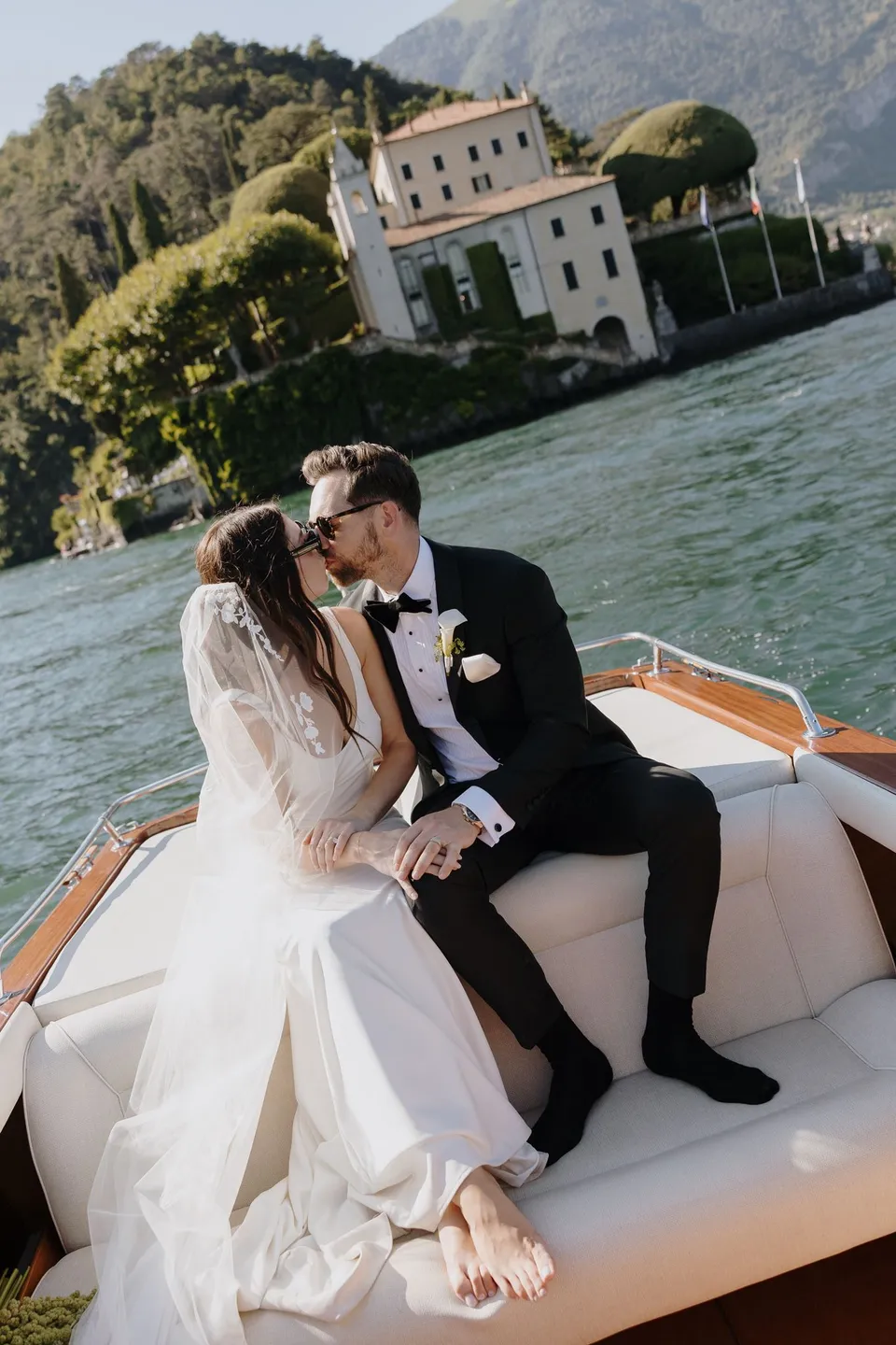 Lyndal and Lachlan kissing on a white boat seat beside a polished timber rail, with a lakeside villa and the boat’s wake behind them, at Lake Como in Lombardy, Italy, for an Elopement Collective elopement. Photograph by George Bowden.