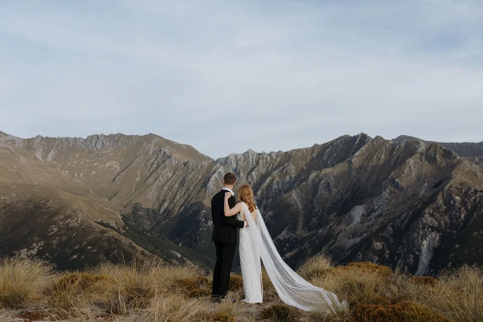 Neve and Hamish kissing among alpine tussock with rugged mountain peaks rising behind them, on Big Geordie above Queenstown, New Zealand, for an Elopement Collective Queenstown elopement. Photo by George Bowden.