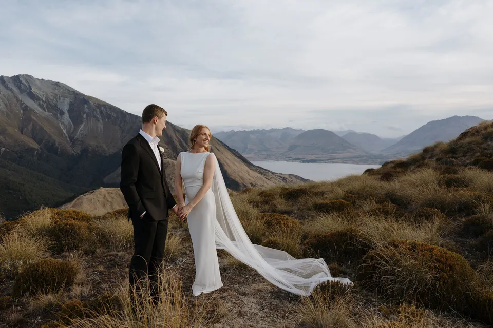 Neve and Hamish holding hands on the mountain ridge, with her cape sweeping across the grass and a lake visible beyond the hills, on Big Geordie above Queenstown, New Zealand, for an Elopement Collective Queenstown elopement. Photo by George Bowden.