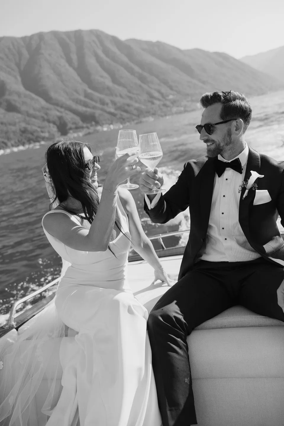 Black and white photograph of Lyndal and Lachlan raising champagne glasses aboard a timber boat, with mountains behind them, at Lake Como in Lombardy, Italy, for an Elopement Collective elopement. Photograph by George Bowden.