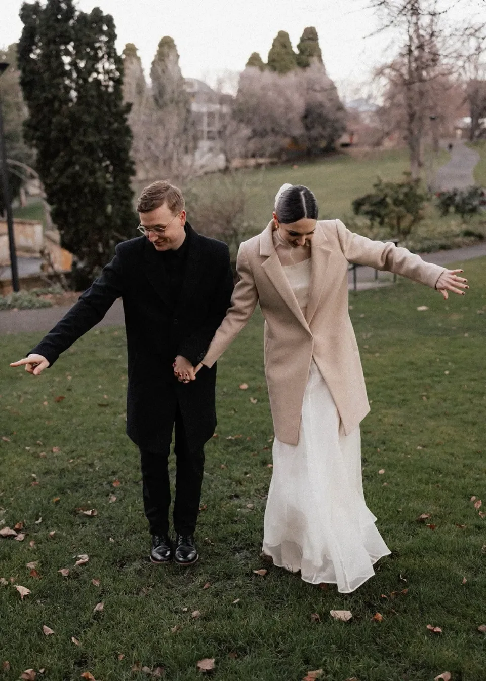 A couple step playfully across a garden lawn with their arms outstretched.