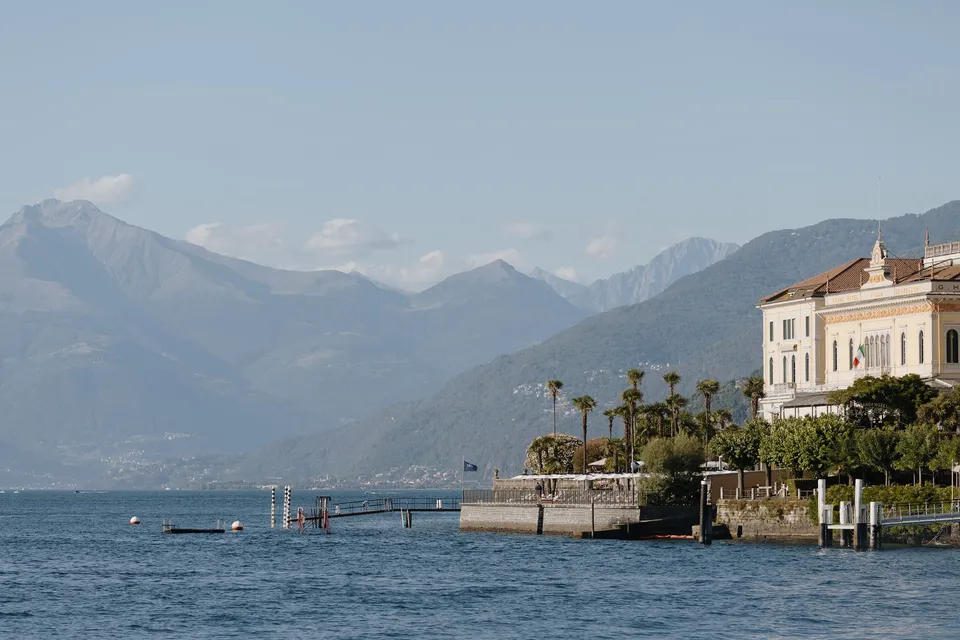 A grand lakeside building, palm trees and steep mountain slopes seen across the blue water, at Lake Como in Lombardy, Italy, for an Elopement Collective elopement. Photograph by George Bowden.