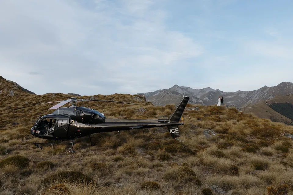 The helicopter resting on an alpine landing site among golden tussock, with mountain ridges beyond, on Big Geordie above Queenstown, New Zealand, for an Elopement Collective Queenstown elopement. Photo by George Bowden.