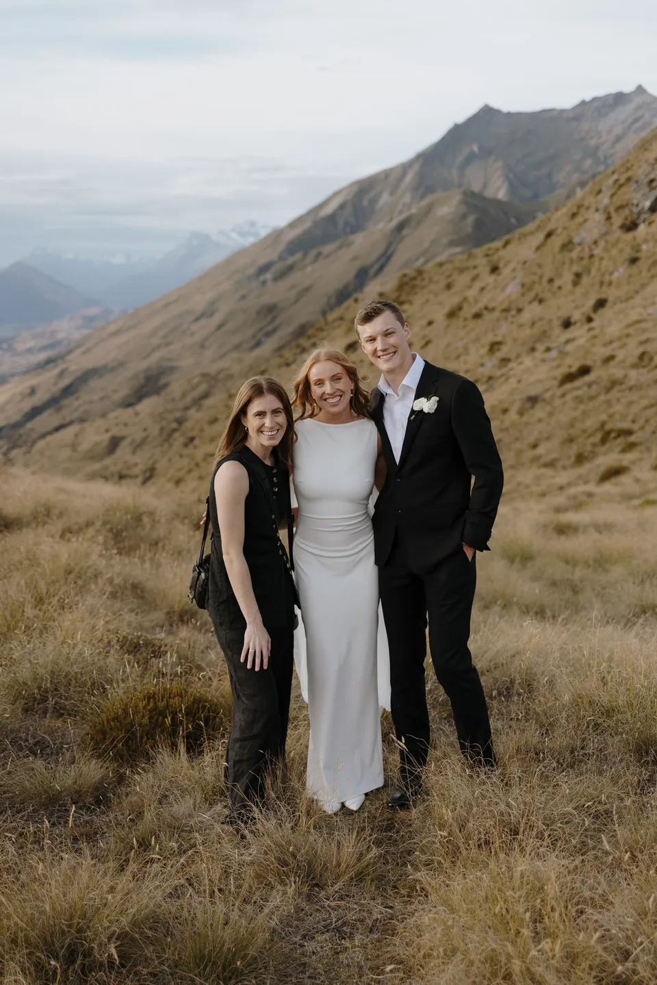 Neve and Hamish smiling with a guest among the golden grasses, with a mountain ridge behind them, on Big Geordie above Queenstown, New Zealand, for an Elopement Collective Queenstown elopement. Photo by George Bowden.