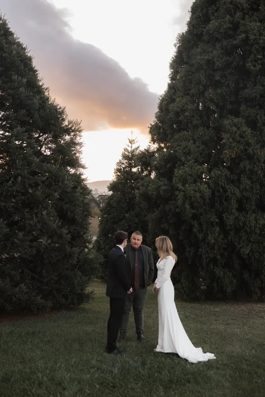 A couple stand beneath tall evergreen trees with warm light in the cloudy sky.