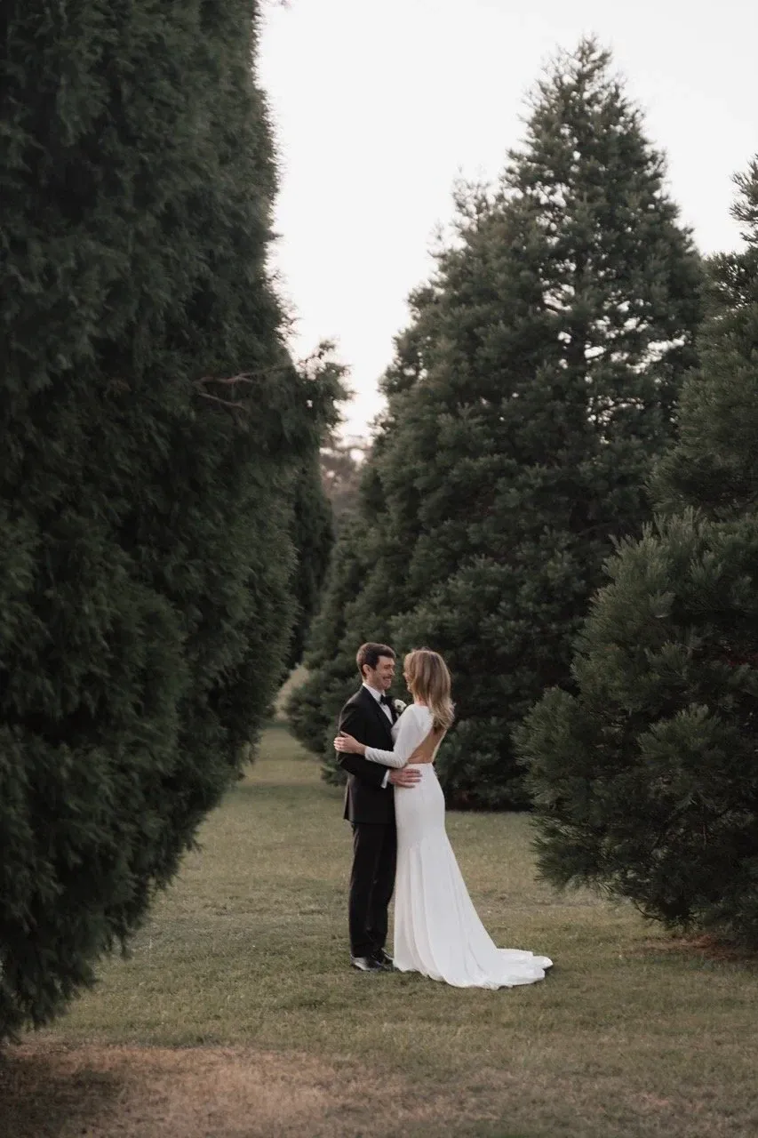 A couple embrace on a grass path between rows of tall evergreen trees.