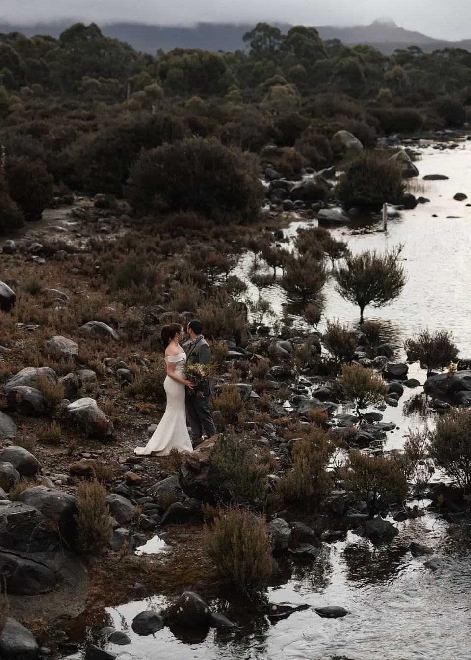 A bride stands on a rocky lakeshore among low shrubs beneath a grey sky.