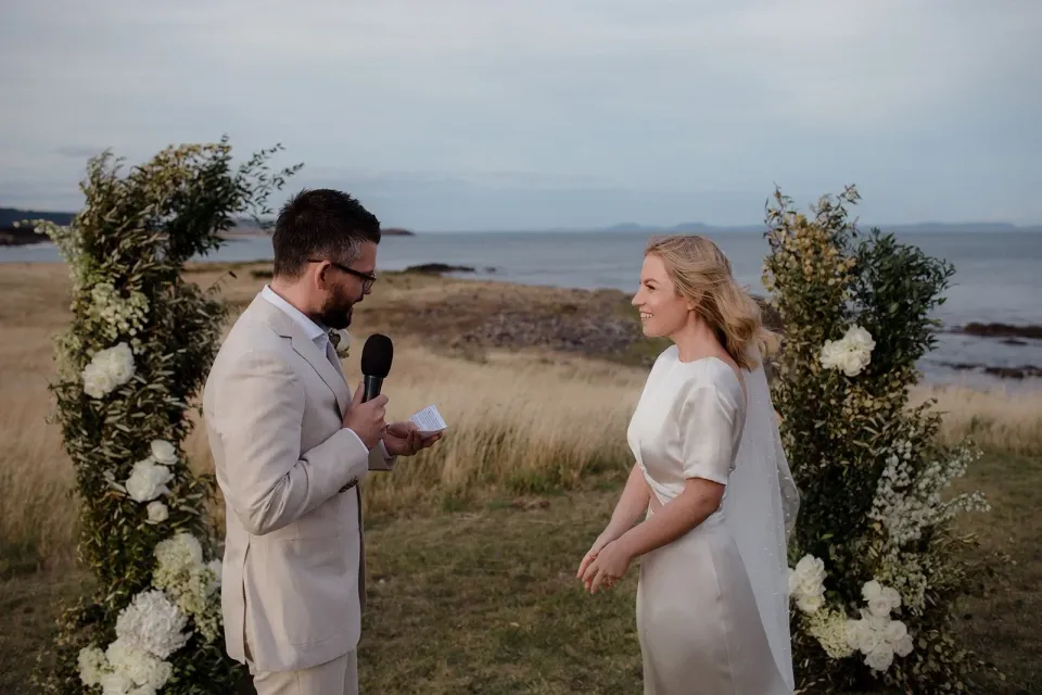 A groom reads his vows into a microphone between floral arrangements overlooking the coast.