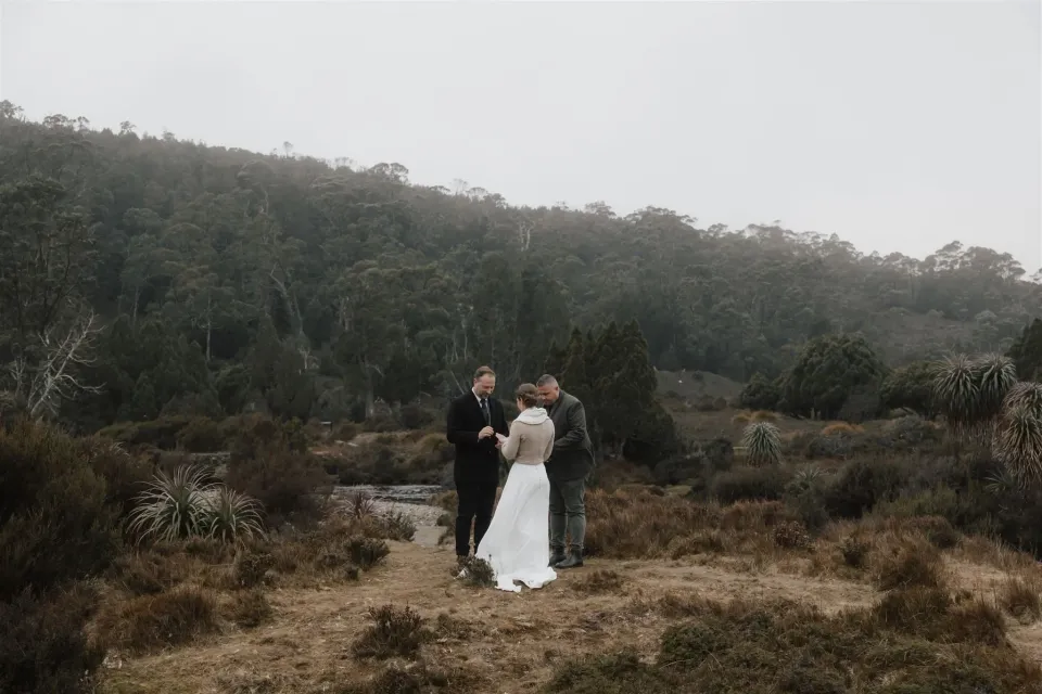 A couple stand together in an open clearing surrounded by forest and scrub.