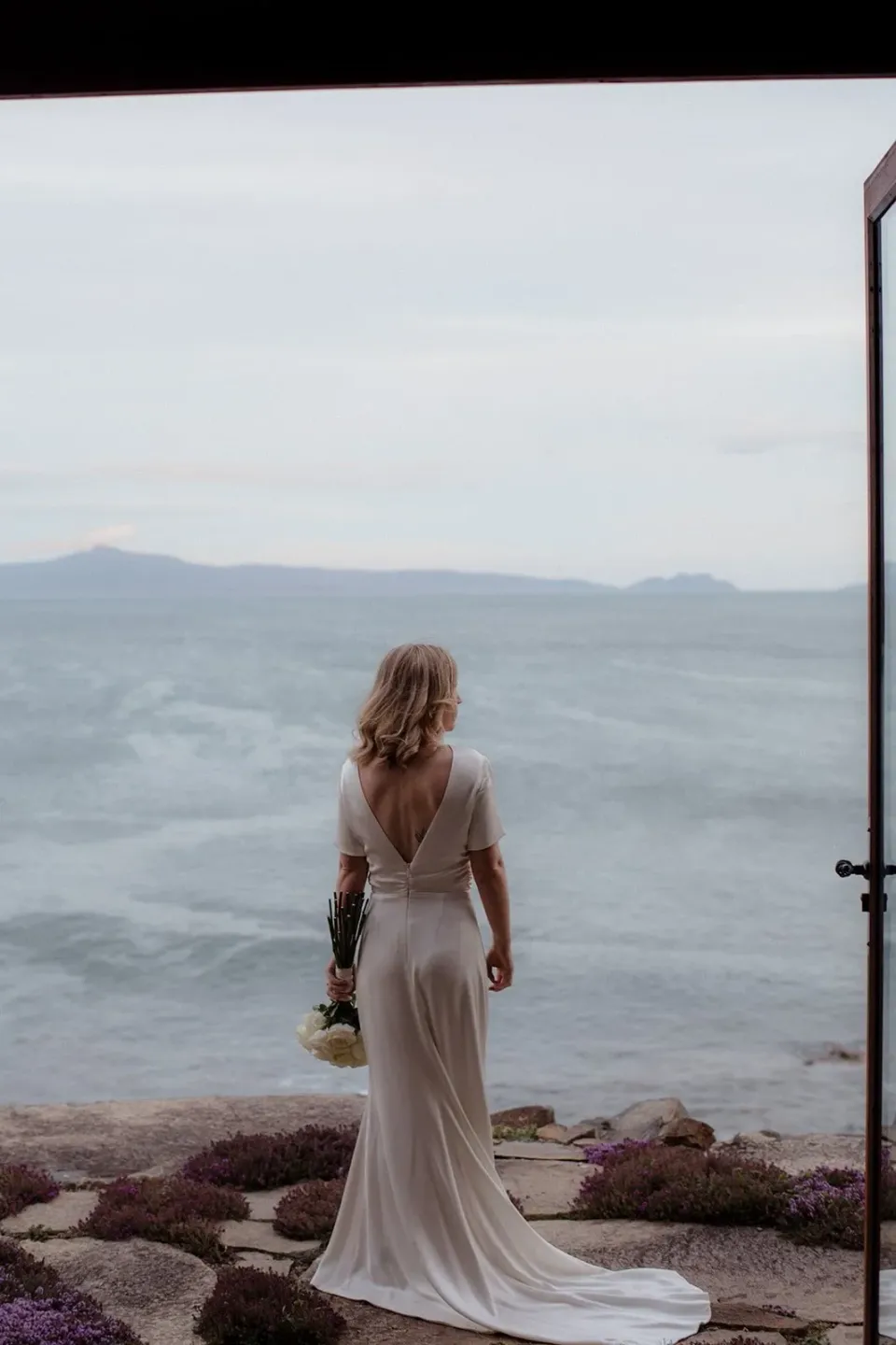 A bride looks out through an open doorway towards a wide lake.