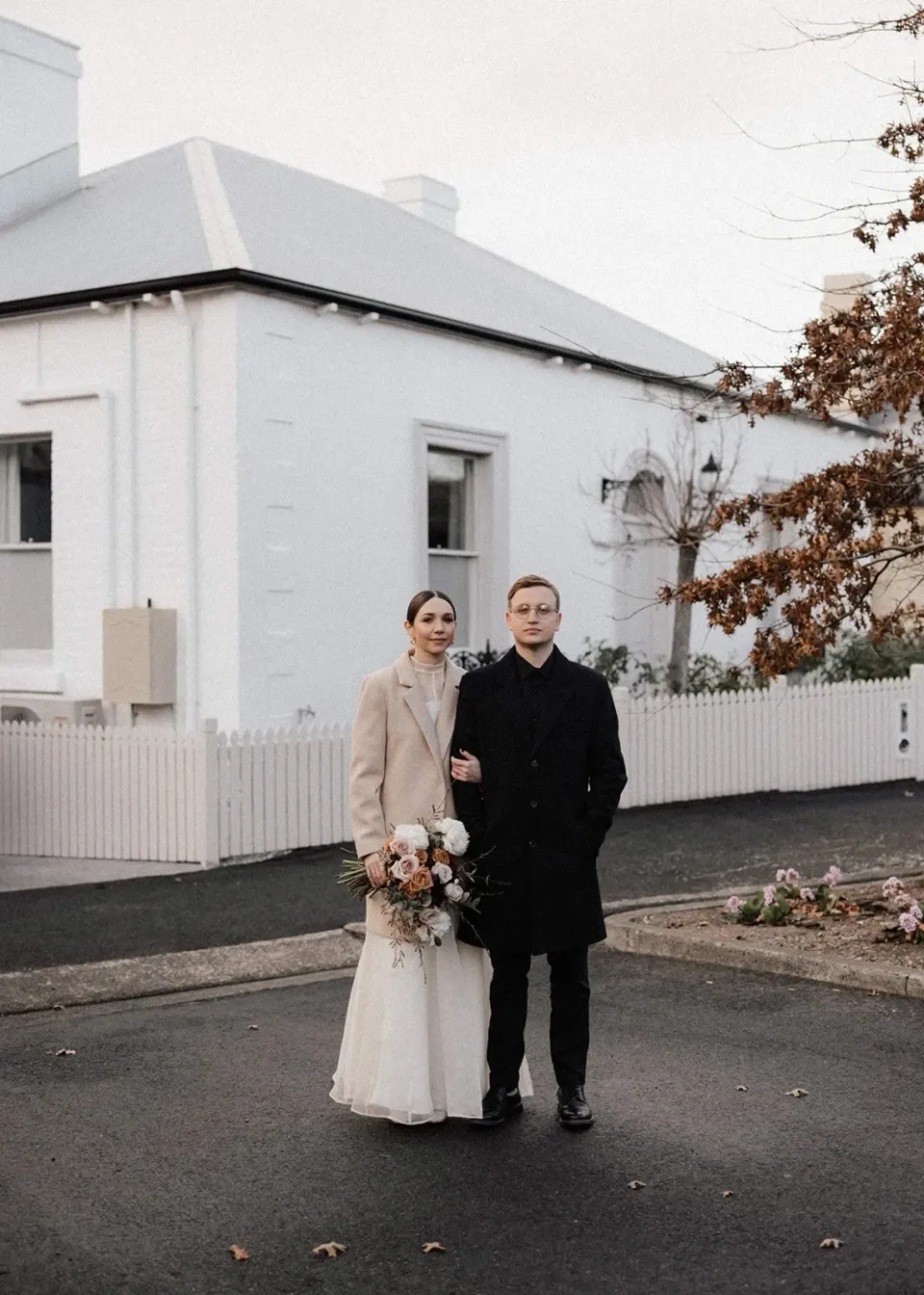 A couple stand beside a pale weatherboard cottage on a Hobart street corner.