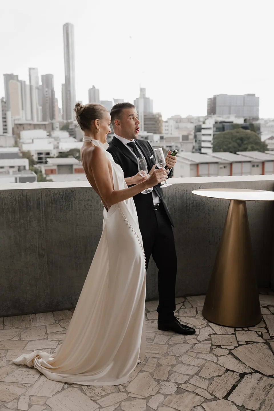 Clare and Matt holding champagne on a terrace with Brisbane’s skyline behind them at The Calile Hotel in Fortitude Valley for their Elopement Collective elopement.