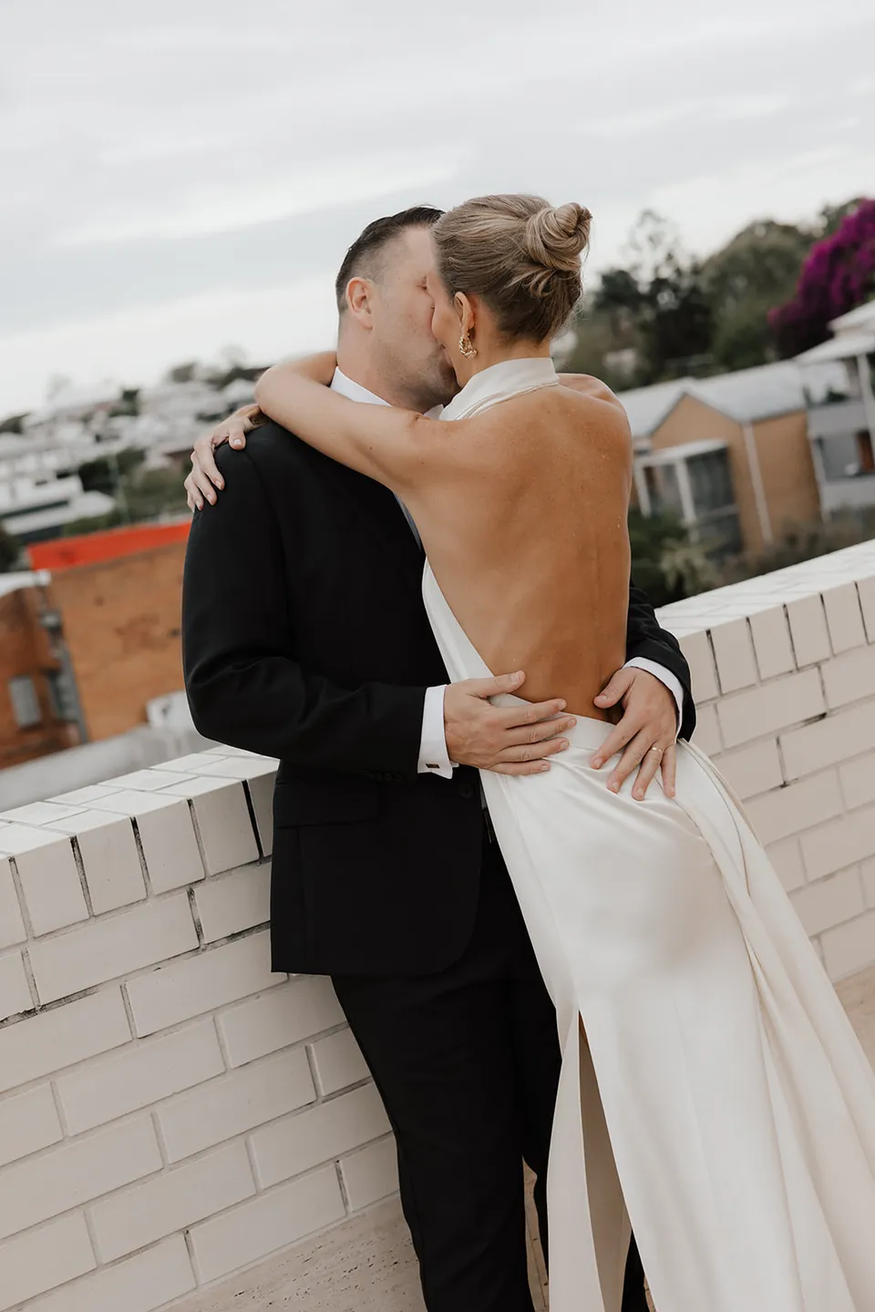 Clare and Matt embracing beside a white brick terrace wall at The Calile Hotel, Fortitude Valley, during their Elopement Collective elopement in Brisbane.
