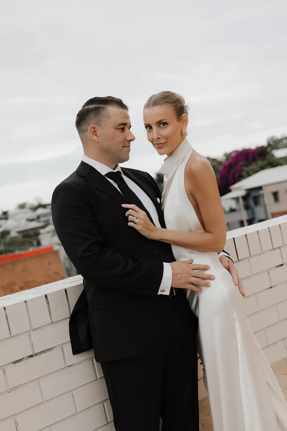 Clare and Matt standing together by a white brick wall with Brisbane behind them at The Calile Hotel, Fortitude Valley, during their Elopement Collective elopement.