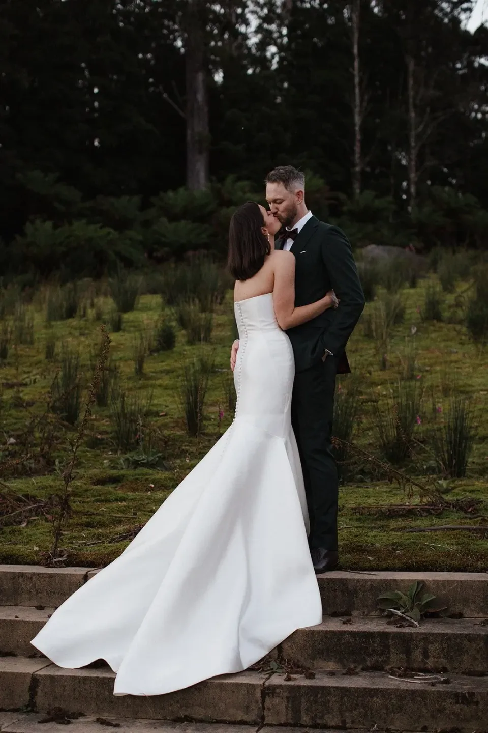 A couple embrace on stone steps beside a forest garden, with the bride's train flowing behind.