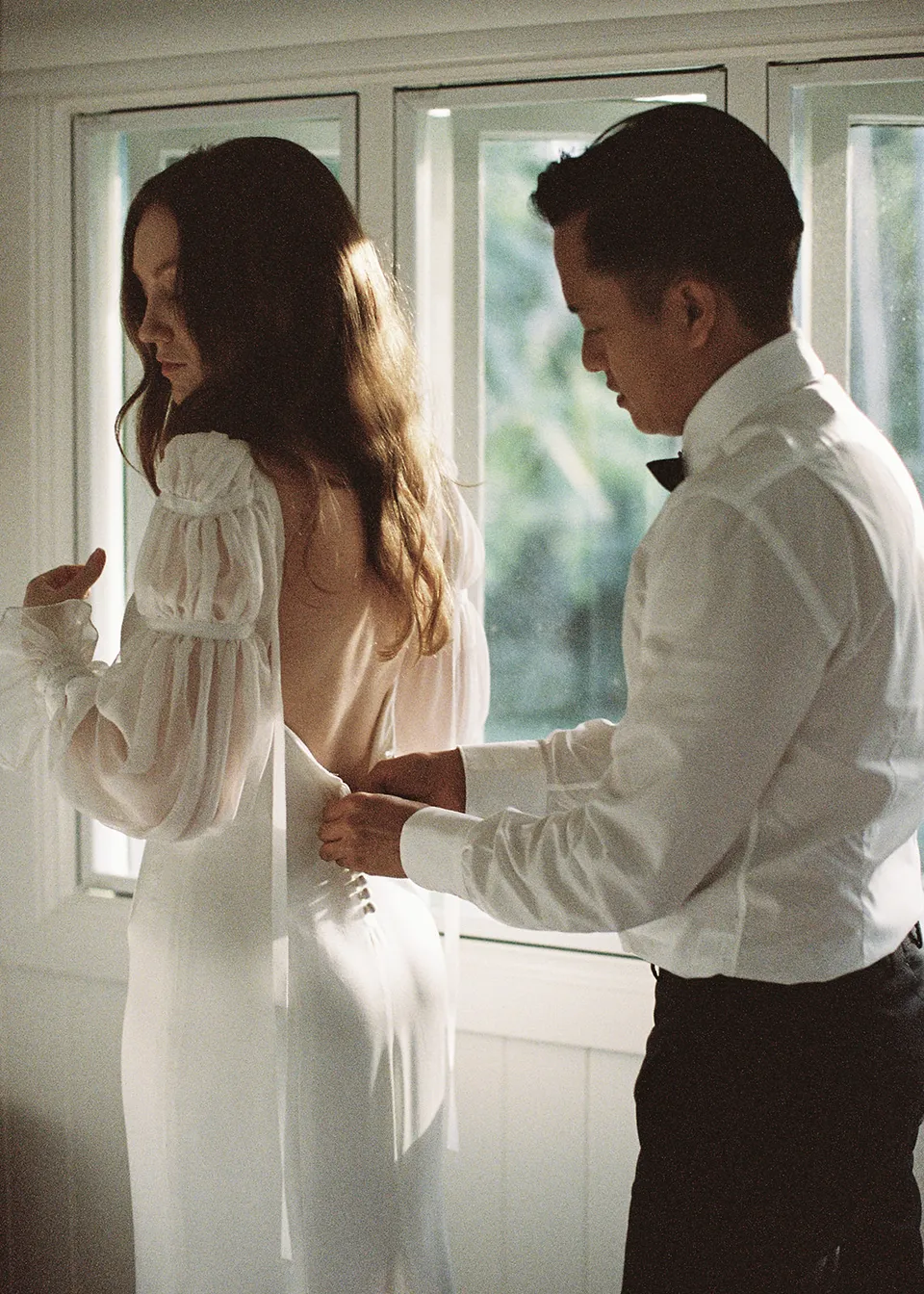 On 35mm film: Elyse looking over her shoulder while Jonathan fastens her dress. At Fig Tree Villa in Ewingsdale, Byron Bay, for an Elopement Collective elopement. Photograph by Bulb Creative.