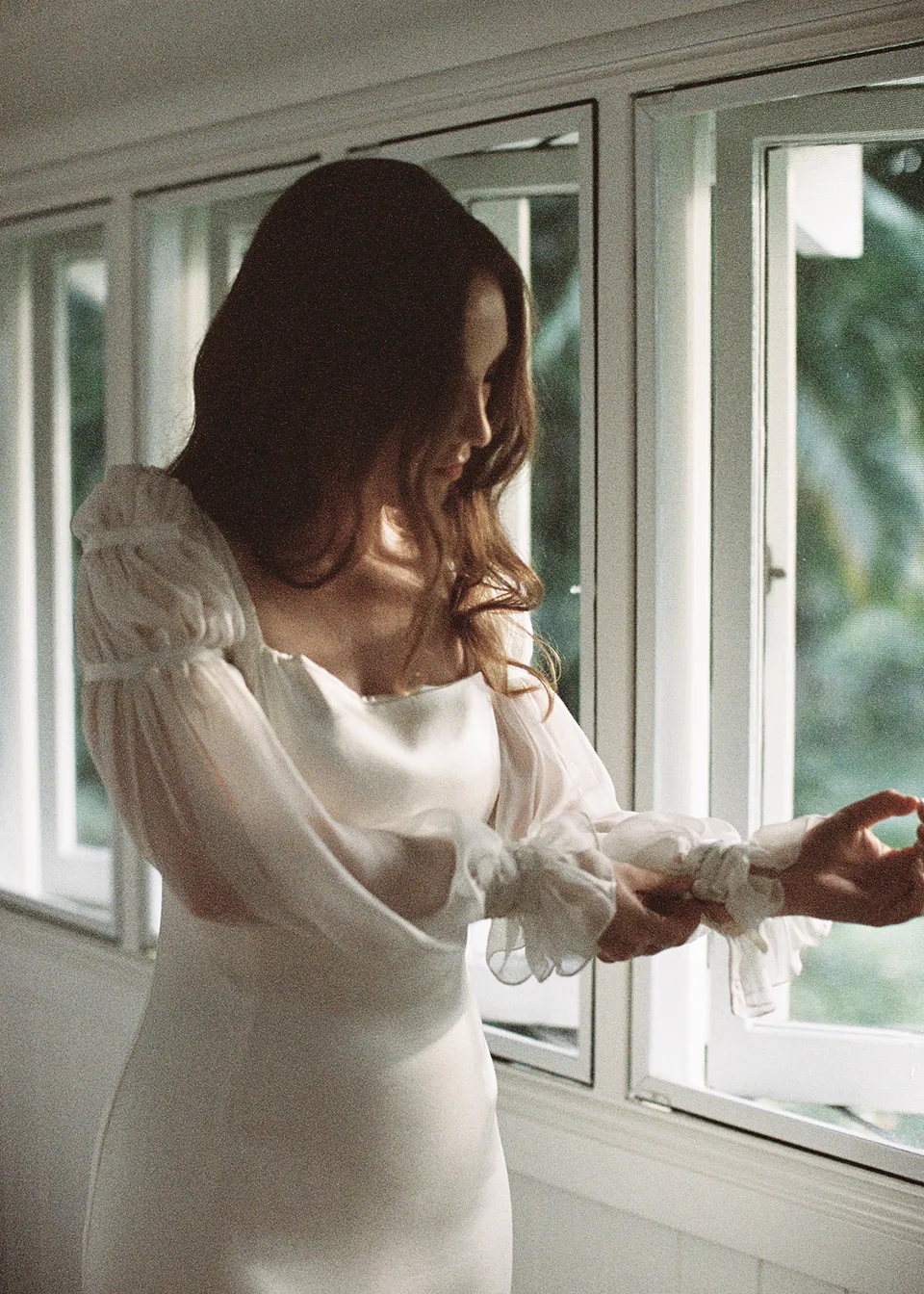 On 35mm film: Elyse adjusting a sheer sleeve beside the bedroom window. At Fig Tree Villa in Ewingsdale, Byron Bay, for an Elopement Collective elopement. Photograph by Bulb Creative.