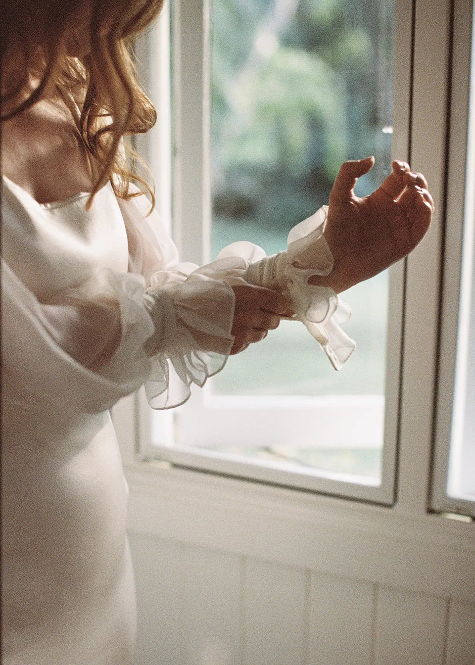 On 35mm film: Close-up of Elyse's gathered sleeve and hands in the window light. At Fig Tree Villa in Ewingsdale, Byron Bay, for an Elopement Collective elopement. Photograph by Bulb Creative.