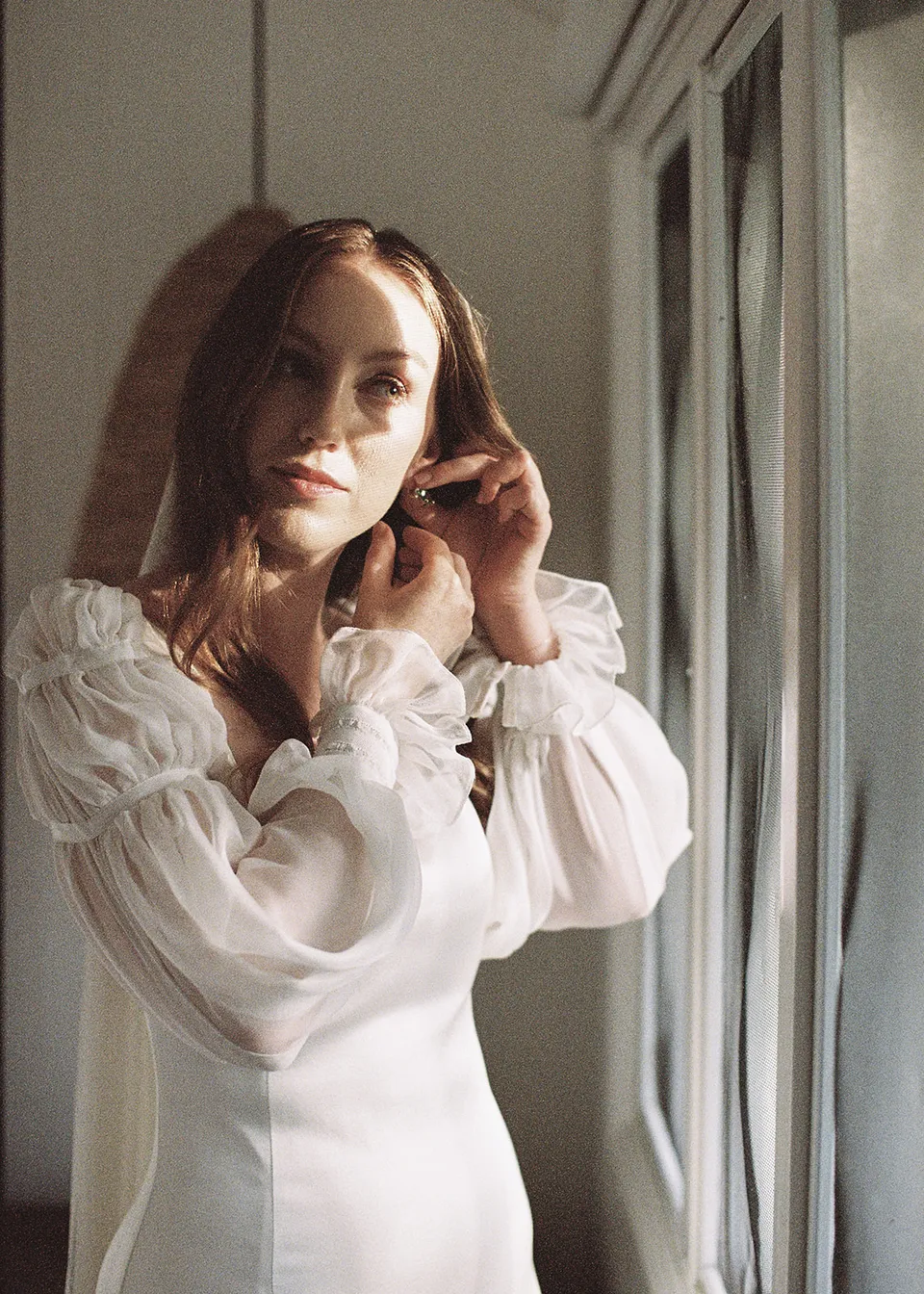 On 35mm film: Elyse fastening her earring beside the window. At Fig Tree Villa in Ewingsdale, Byron Bay, for an Elopement Collective elopement. Photograph by Bulb Creative.