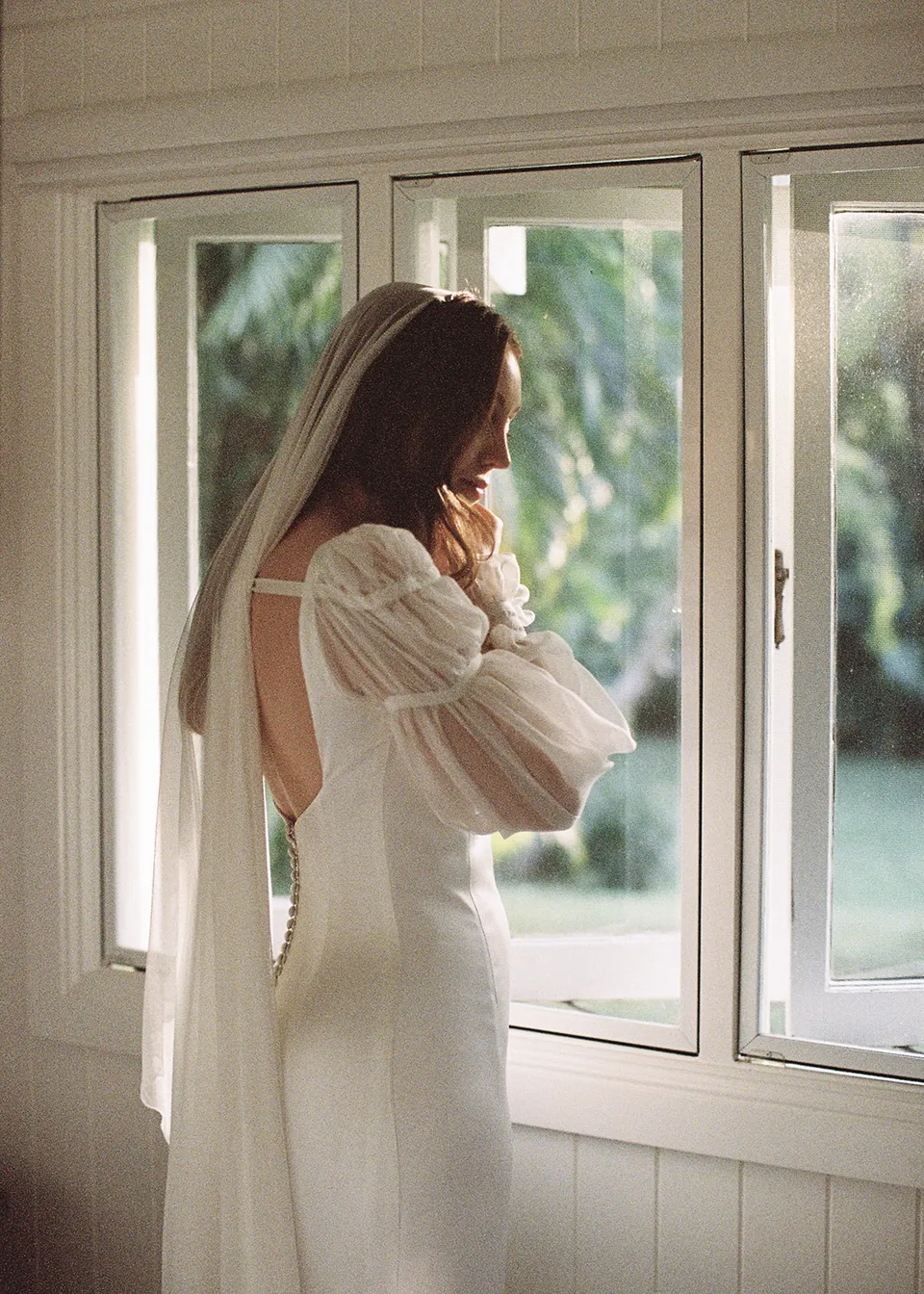 On 35mm film: Rear view of Elyse beneath her veil beside the open window. At Fig Tree Villa in Ewingsdale, Byron Bay, for an Elopement Collective elopement. Photograph by Bulb Creative.