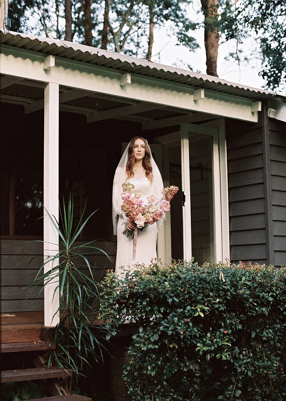 On 35mm film: Elyse standing on the cottage verandah with her bouquet, with flowers by The Perfect Party Co. At Fig Tree Villa in Ewingsdale, Byron Bay, for an Elopement Collective elopement. Photograph by Bulb Creative.