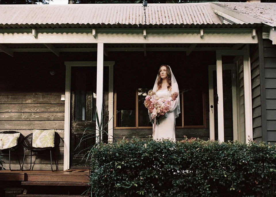 On 35mm film: Wide portrait of Elyse on the verandah of the weatherboard cottage. At Fig Tree Villa in Ewingsdale, Byron Bay, for an Elopement Collective elopement. Photograph by Bulb Creative.