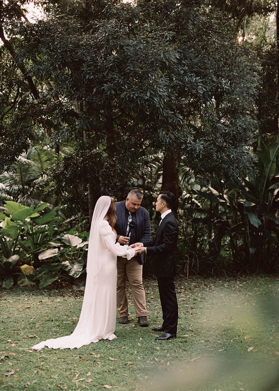 On 35mm film: Elyse and Jonathan holding hands with celebrant Josh Withers on the garden lawn. At Fig Tree Villa in Ewingsdale, Byron Bay, for an Elopement Collective elopement. Photograph by Bulb Creative.