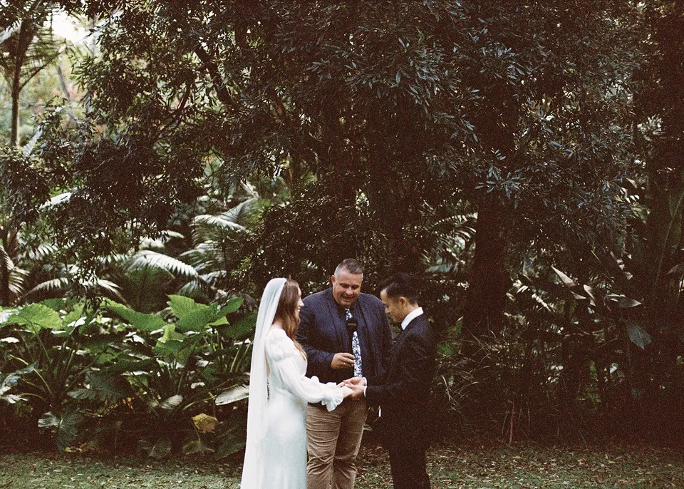 On 35mm film: Wide view of the small ceremony framed by lush garden foliage. At Fig Tree Villa in Ewingsdale, Byron Bay, for an Elopement Collective elopement. Photograph by Bulb Creative.