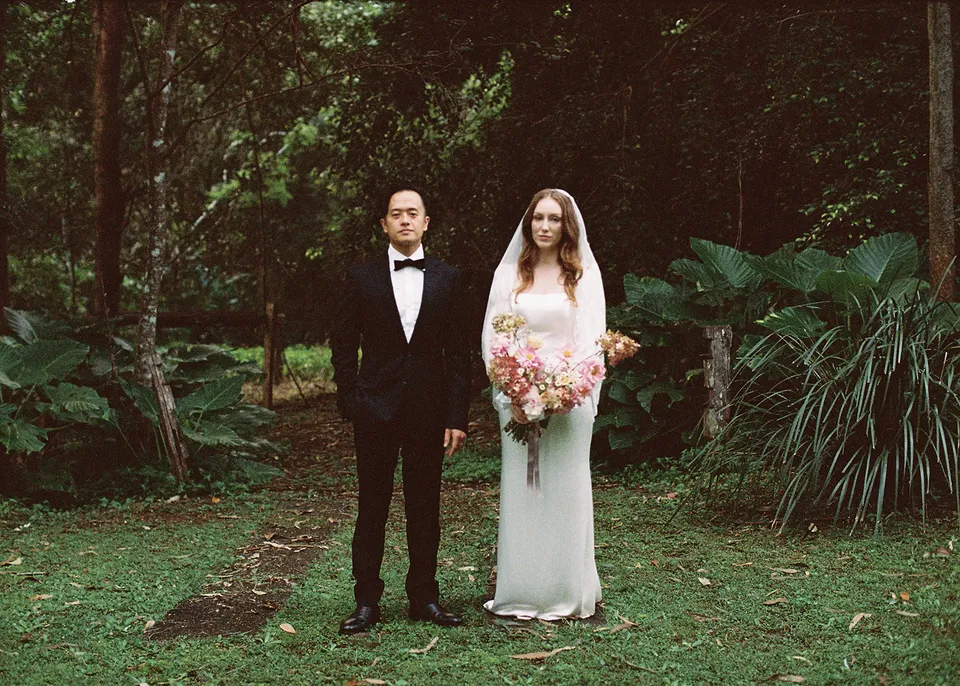 On 35mm film: garden portrait of the couple with Elyse's bouquet, with flowers by The Perfect Party Co. At Fig Tree Villa in Ewingsdale, Byron Bay, for an Elopement Collective elopement. Photograph by Bulb Creative.