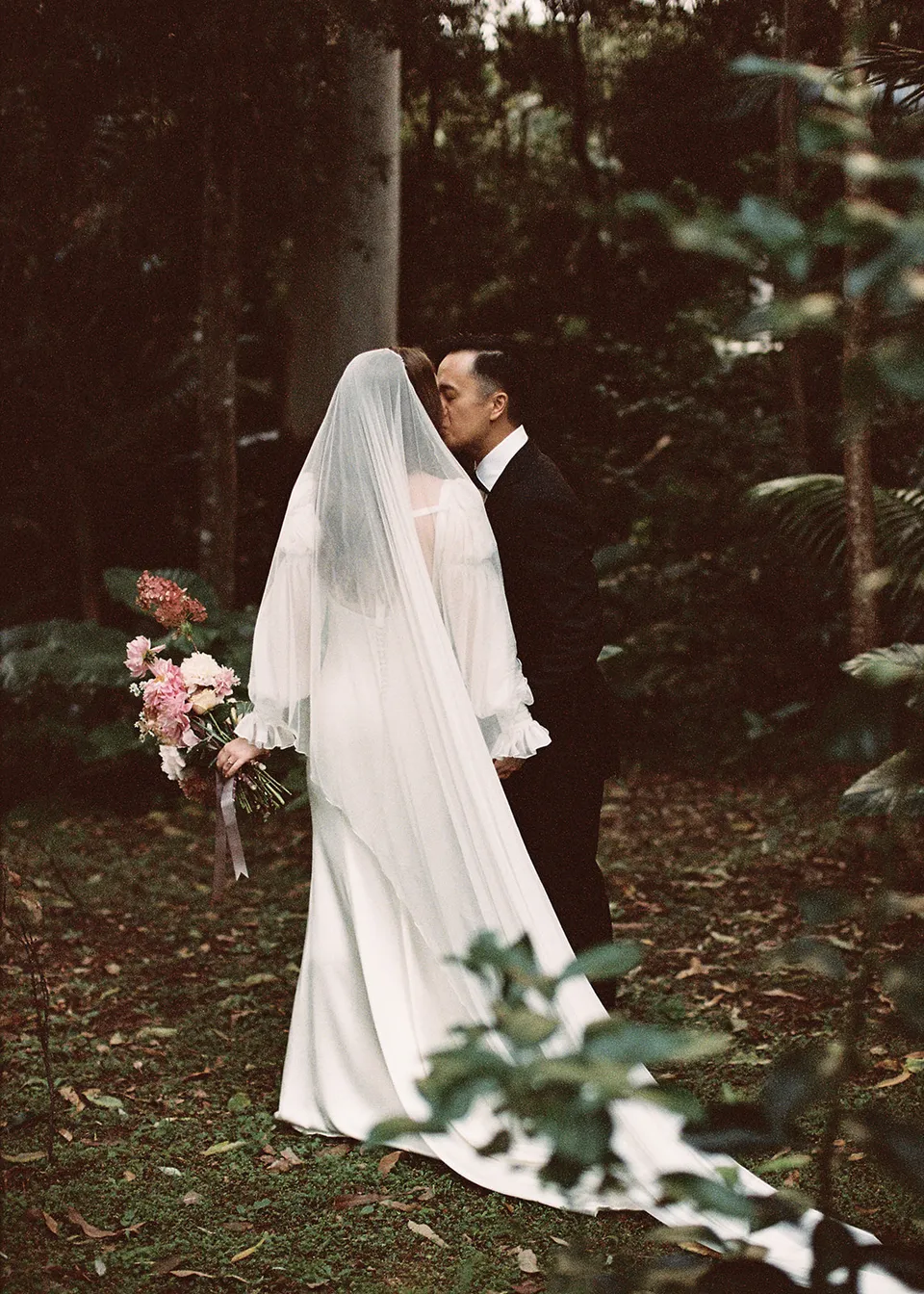 On 35mm film: Jonathan kissing Elyse's cheek beside the trees. At Fig Tree Villa in Ewingsdale, Byron Bay, for an Elopement Collective elopement. Photograph by Bulb Creative.