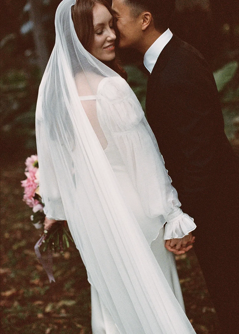 On 35mm film: Elyse smiling with her eyes closed as Jonathan kisses her cheek. At Fig Tree Villa in Ewingsdale, Byron Bay, for an Elopement Collective elopement. Photograph by Bulb Creative.